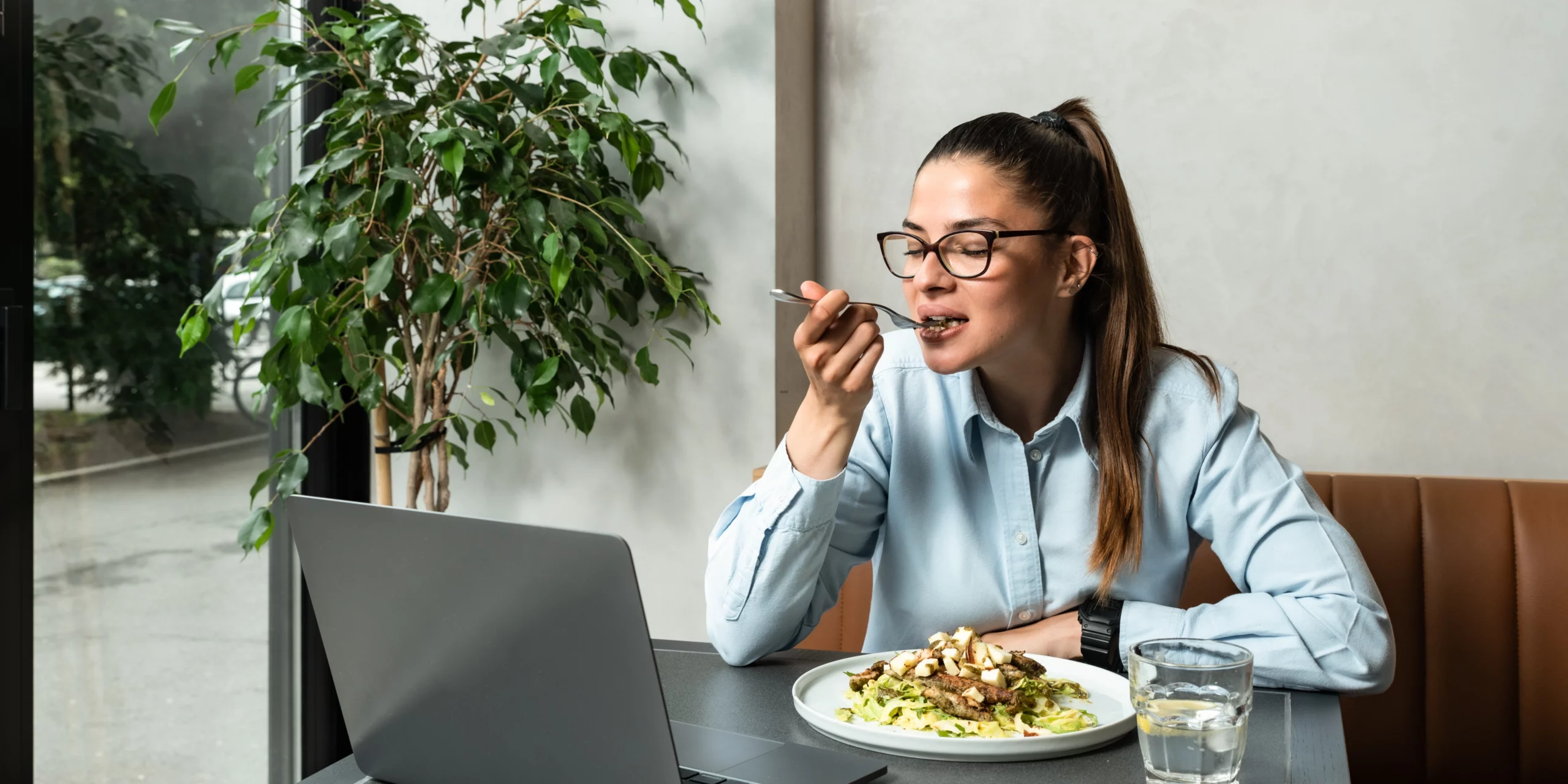 Une femme concentrée mange son repas tout en travaillant sur son ordinateur portable, lumière naturelle douce.</p>
<p>