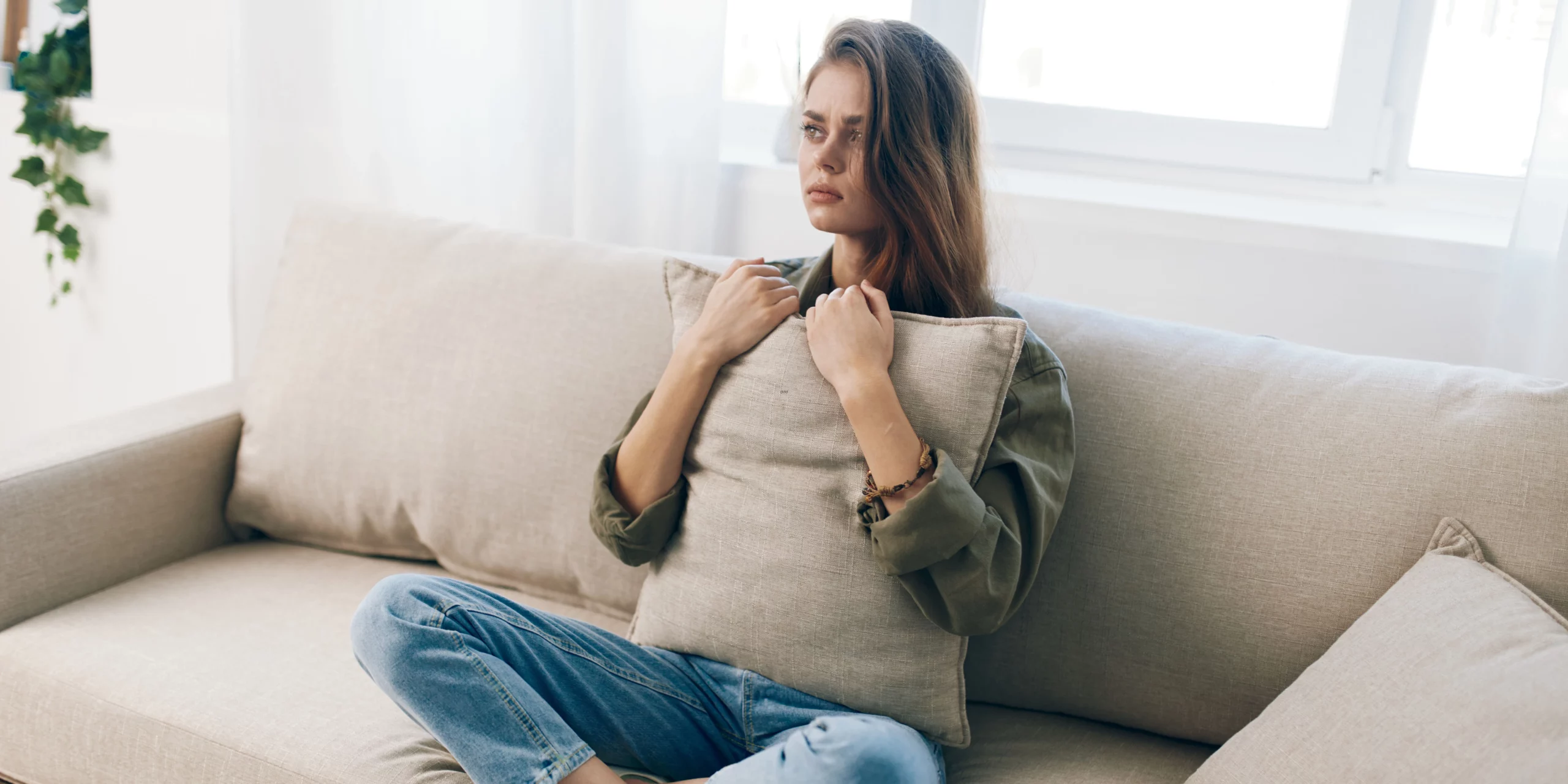Une femme assise sur un fauteuil, serrant un coussin contre elle, le regard inquiet et le corps tendu par l’anxiété.