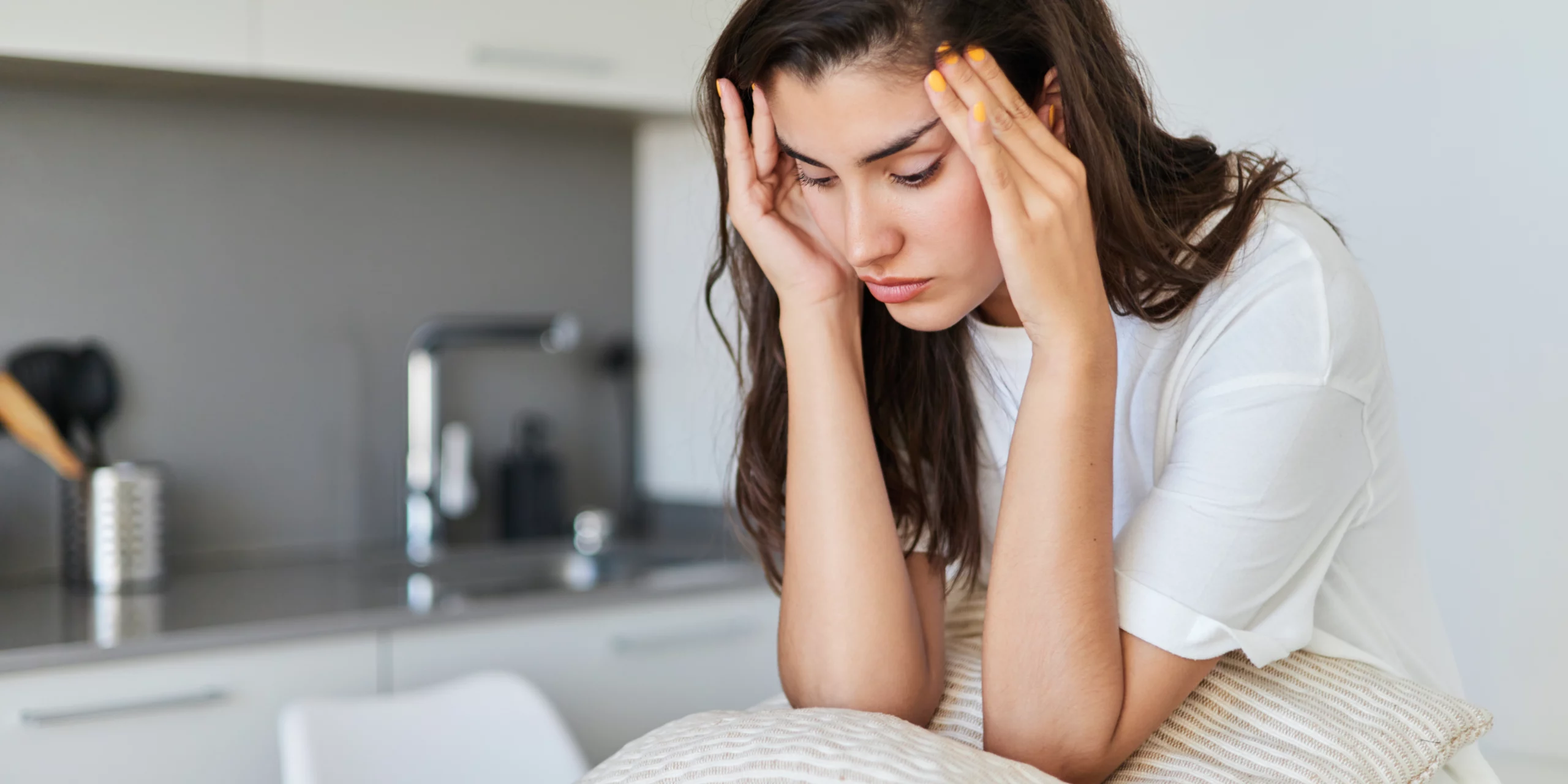 Une jeune femme, l'air anxieux et fatigué, se tient la tête à deux mains, les coudes posés sur un coussin. Elle semble submergée par le stress ou une forte douleur, son regard perdu vers le bas. Le décor est celui d'une cuisine moderne et claire, mais son expression contraste avec le calme de la pièce, évoquant la charge mentale ou l'épuisement émotionnel.
