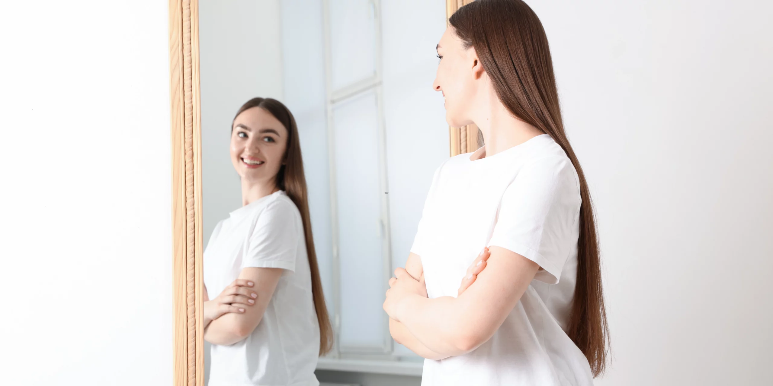 Une jeune femme rayonnante, aux longs cheveux bruns soyeux, contemple son reflet avec une expression de profonde satisfaction personnelle. Debout face à un miroir au cadre en bois clair, elle affiche un sourire serein et une posture assurée, les bras croisés. La scène se déroule dans un intérieur baigné de lumière naturelle, symbolisant un moment de reconnexion à soi et d'épanouissement intérieur dans un cadre doux et minimaliste.