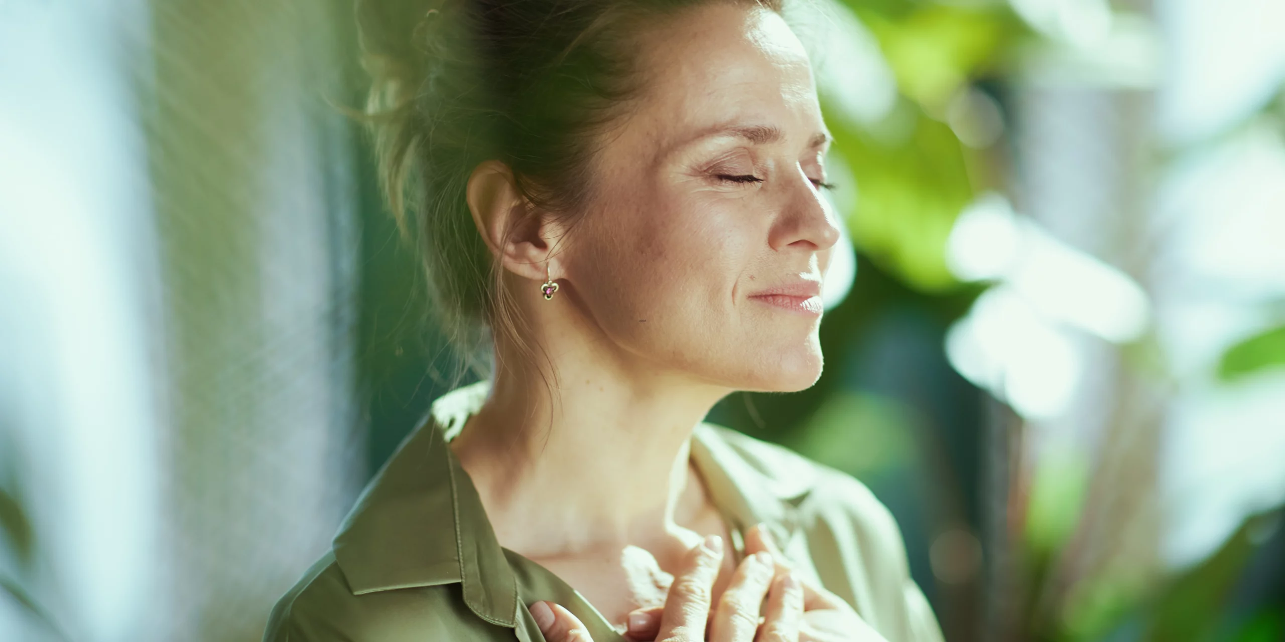Une femme aux yeux clos et au sourire serein, les mains posées sur le cœur, baignée dans une lumière naturelle douce et verdoyante. Elle semble prendre une grande inspiration de bien-être, loin du stress des notifications.