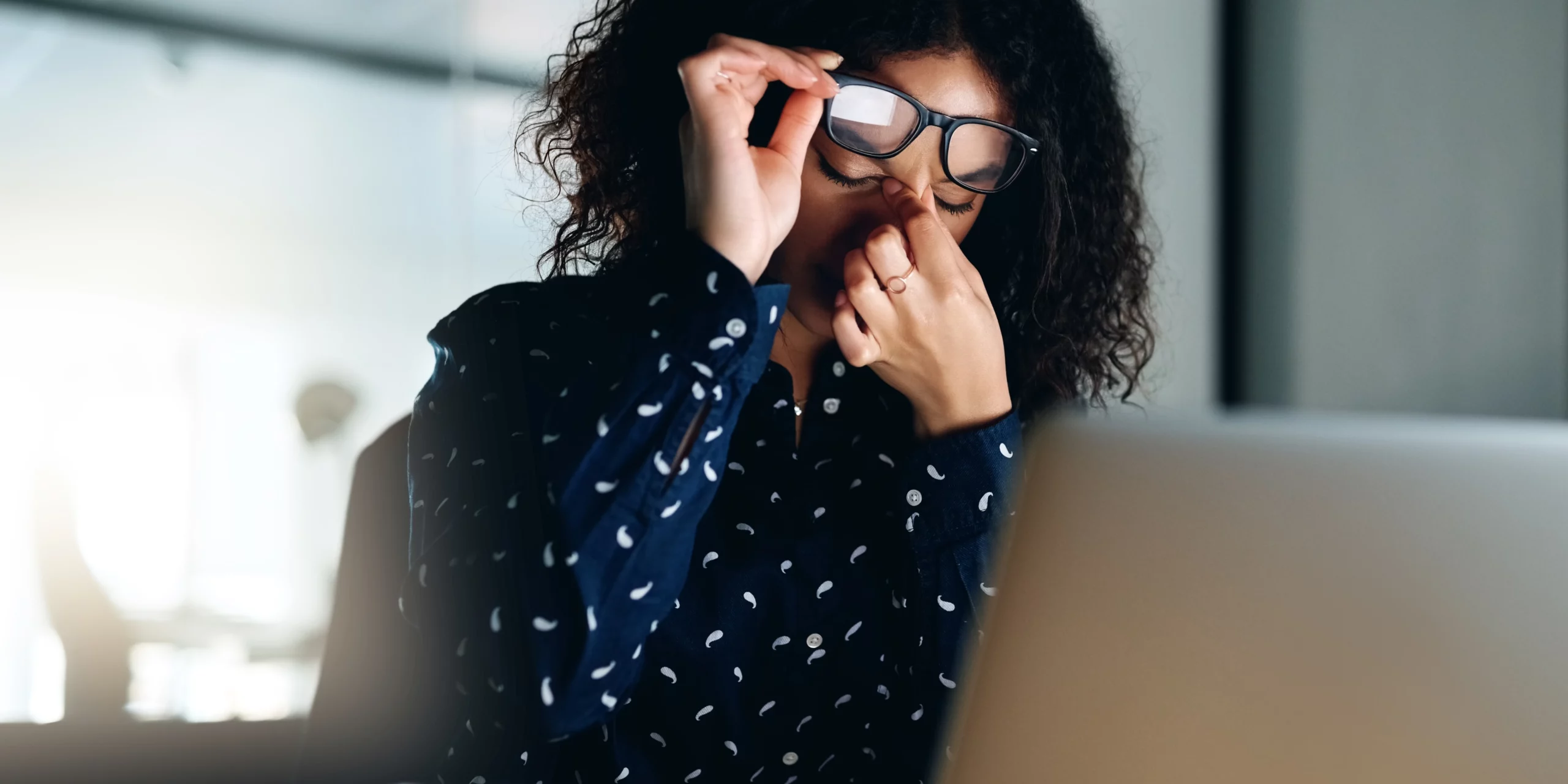 Une femme devant son ordinateur soulève ses lunettes pour se masser l'arête du nez, les yeux fermés. Elle affiche un air de fatigue ou de concentration intense, illustrant parfaitement le besoin de faire une pause loin des écrans.